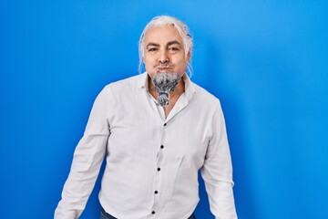 Middle age man with grey hair standing over blue background puffing cheeks with funny face. mouth inflated with air, crazy expression.