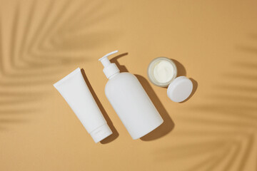 Skin care beauty concept. Flat lay photo of white cosmetics bottles, cream jar and tropical leaves shadow on sandy background. Summer cosmetics mockup idea.