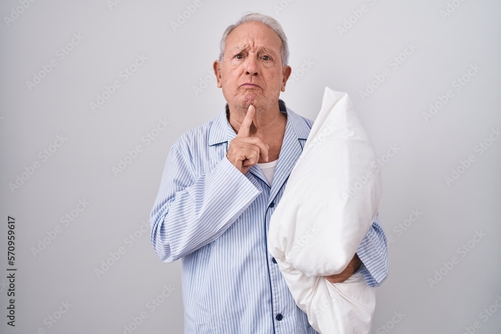 Poster senior man with grey hair wearing pijama hugging pillow thinking concentrated about doubt with finge