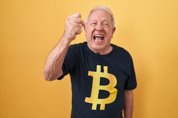 Senior man with grey hair wearing bitcoin t shirt angry and mad raising fist frustrated and furious...