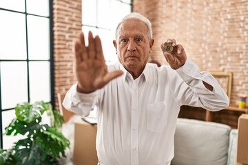 Senior man with grey hair holding virtual currency bitcoin with open hand doing stop sign with...