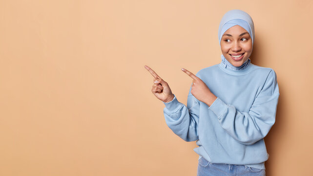 Positive Islamic woman wears blue hijab and jumper points index fingers aside demonstrates copy space for your promotional content has happy expression isolated over brown backgground. Look at this