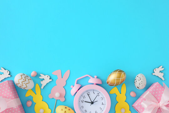 Easter party concept. Flat lay photo of color eggs, cute rabbits, gift boxes and pink alarm clock on pastel blue background with copyspace. Invitation holiday card idea.