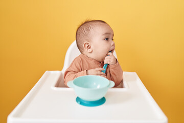 Adorable hispanic toddler sitting on highchair eating over isolated yellow background