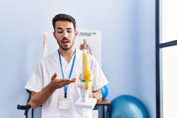 Young hispanic man wearing physiotherapist uniform holding anatomical model of knee at rehab clinic