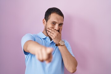 Hispanic man standing over pink background laughing at you, pointing finger to the camera with hand...