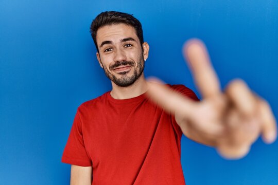 Young hispanic man with beard wearing red t shirt over blue background smiling looking to the camera showing fingers doing victory sign. number two.