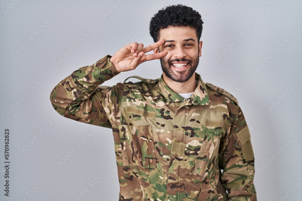 Sticker Arab man wearing camouflage army uniform doing peace symbol with fingers over face, smiling cheerful showing victory