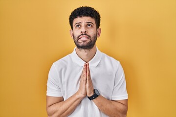 Arab man standing over yellow background begging and praying with hands together with hope...