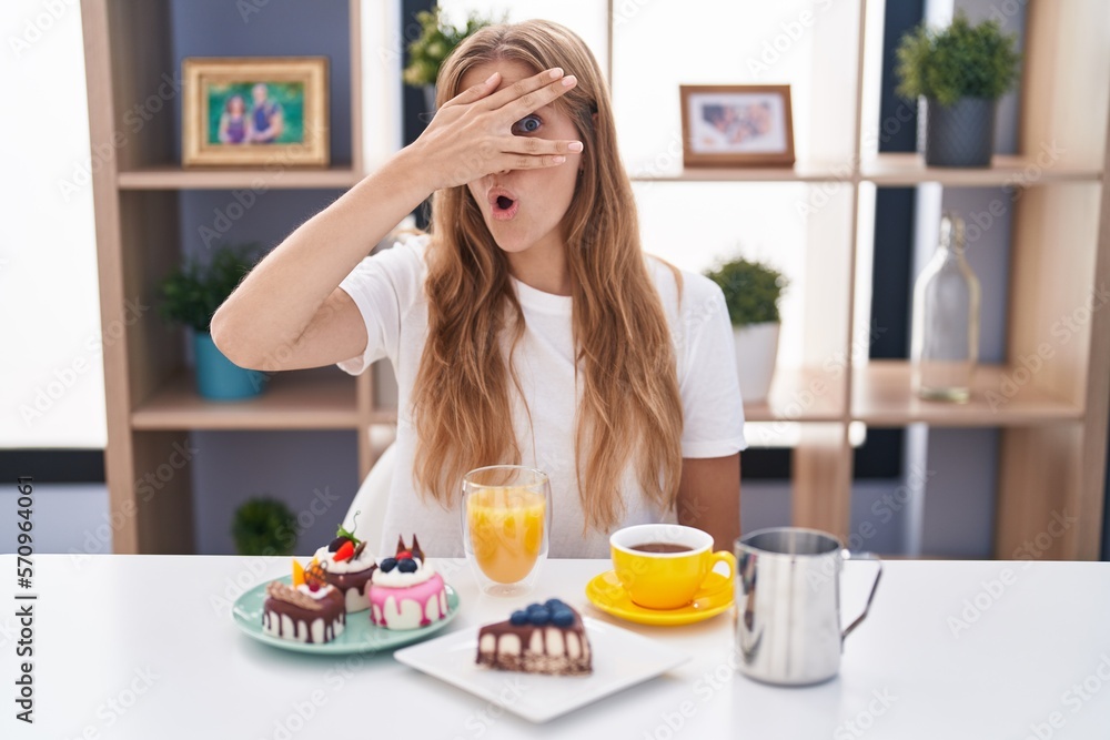 Sticker young caucasian woman eating pastries t for breakfast peeking in shock covering face and eyes with h