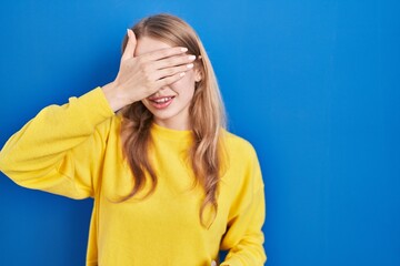 Young caucasian woman standing over blue background smiling and laughing with hand on face covering eyes for surprise. blind concept.