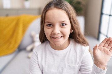 Adorable hispanic girl sitting on sofa saying hello with hand at home