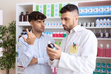 Two hispanic men pharmacist speaking to client showing medication label bottle at pharmacy