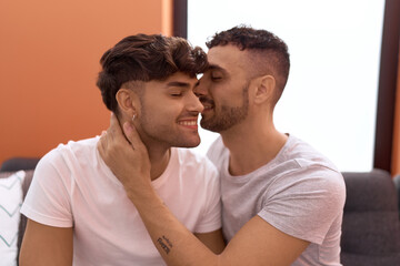 Two hispanic men couple sitting on sofa hugging each other and kissing at home