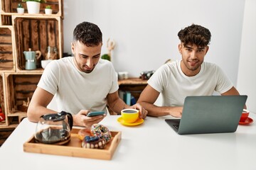 Fototapeta na wymiar Two hispanic men couple having breakfast using smartphone and laptop at home