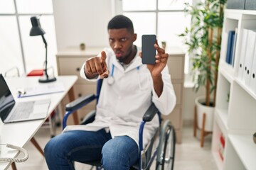 African american doctor man sitting on wheelchair holding smartphone pointing with finger to the camera and to you, confident gesture looking serious