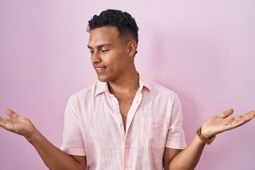 Young hispanic man standing over pink background smiling showing both hands open palms, presenting and advertising comparison and balance