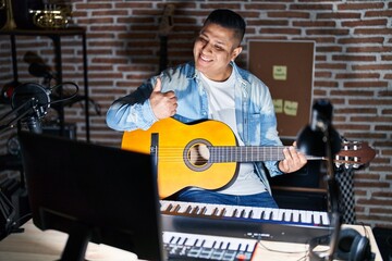 Hispanic young man playing classic guitar at music studio doing happy thumbs up gesture with hand. approving expression looking at the camera showing success.