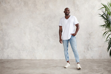 Handsome African American young man stands full body, looks at camera, against bright textured wall in studio. Male model wears casual clothes white T-shirt and jeans © Anna Zhukkova