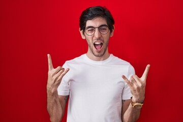 Naklejka premium Young hispanic man standing over red background shouting with crazy expression doing rock symbol with hands up. music star. heavy music concept.