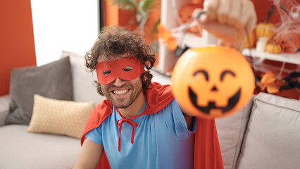 Young hispanic man wearing superhero costume holding halloween pumpkin basket at home