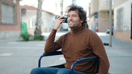 Young hispanic man listening voice message by smartphone sitting on wheelchair at street