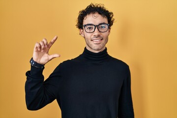 Hispanic man standing over yellow background smiling and confident gesturing with hand doing small size sign with fingers looking and the camera. measure concept.