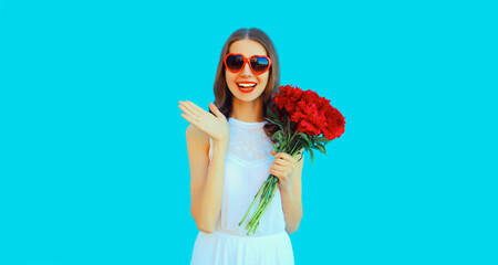 Portrait of beautiful smiling woman with bouquet of red rose flowers in heart shaped sunglasses on blue background