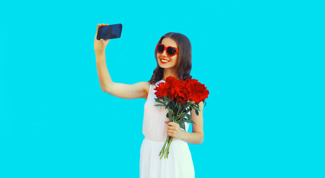 Portrait of beautiful smiling woman taking selfie with phone holding bouquet of red rose flowers in heart shaped sunglasses on blue background