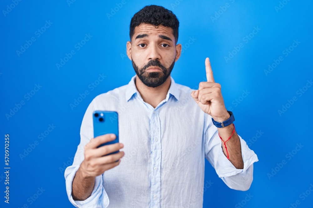 Wall mural Hispanic man with beard using smartphone typing message pointing up looking sad and upset, indicating direction with fingers, unhappy and depressed.