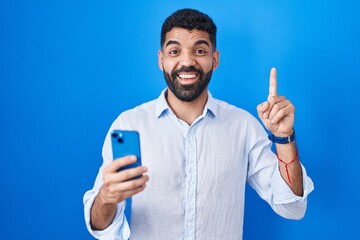 Hispanic man with beard using smartphone typing message smiling amazed and surprised and pointing up with fingers and raised arms.