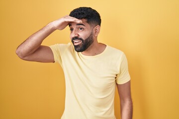 Hispanic man with beard standing over yellow background very happy and smiling looking far away with hand over head. searching concept.