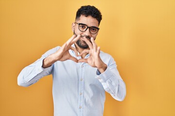 Hispanic man with beard standing over yellow background smiling in love doing heart symbol shape with hands. romantic concept.