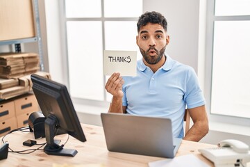 Hispanic man with beard working at the office with laptop holding thanks banner scared and amazed with open mouth for surprise, disbelief face