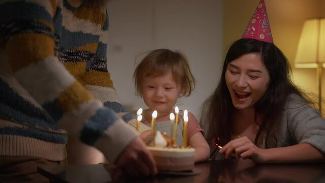 Small family celebrating son birthday at home. Dad and mom whistle a pipe smile and clap hands. Excited little boy looking at cake with burning candles. Happy family clapping hands together. Kid wish