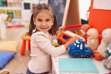Adorable hispanic girl playing with truck toy sitting on floor at kindergarten
