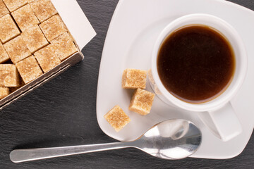 A few cubes of brown sugar on a white saucer with a cup of coffee on a slate stone, macro, top view.