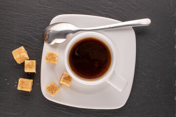 A few cubes of brown sugar on a white saucer with a cup of coffee on a slate stone, macro, top view.