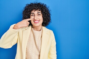 Fototapeta na wymiar Young brunette woman with curly hair standing over blue background pointing with hand finger to face and nose, smiling cheerful. beauty concept