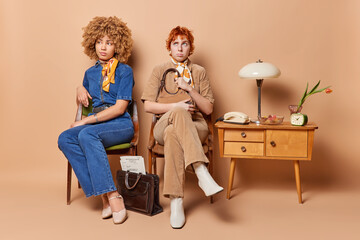 Indoor shot of pensive two women from 90s sit next to each other on chair wear stylish clothes pose...