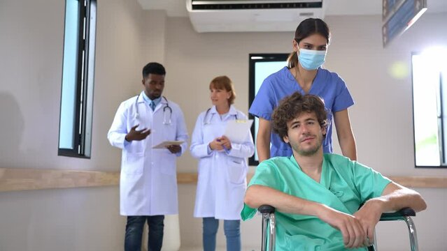 Selective focus of young Caucasian female nurse in a face mask pushing young Caucasian male patient sitting in wheelchair at hospital corridor with blurred two doctors walking discussing in background