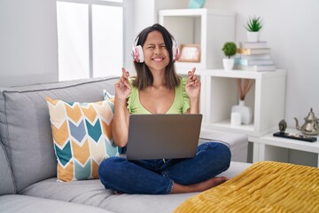 Hispanic young woman using laptop at home gesturing finger crossed smiling with hope and eyes closed. luck and superstitious concept.