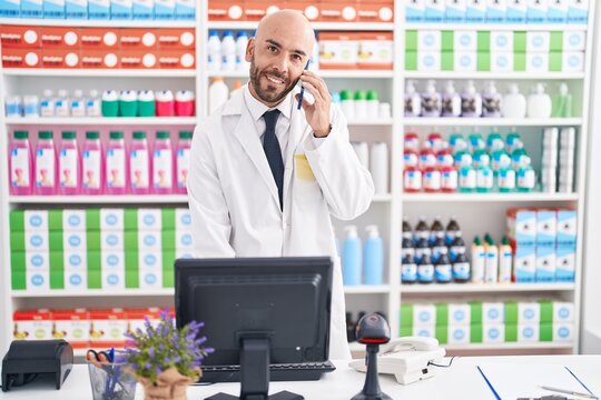 Young bald man pharmacist talking on smartphone using computer at pharmacy
