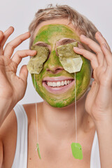 Close up portrait of happy short haired young woman smiles broadly holds tea bags on eyes for skin...
