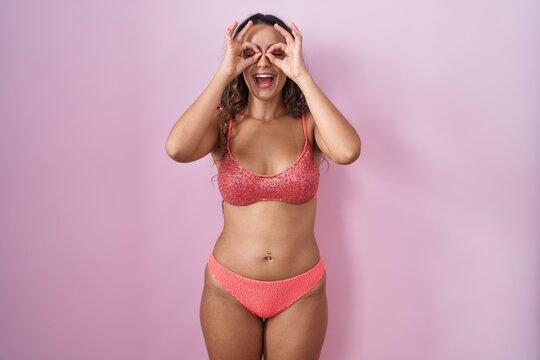 Young hispanic woman wearing lingerie over pink background doing ok gesture like binoculars sticking tongue out, eyes looking through fingers. crazy expression.
