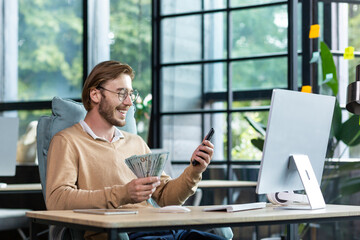 A happy young man is sitting at the table in the lobby, working at the computer. Holds cash, dollars and uses the phone.