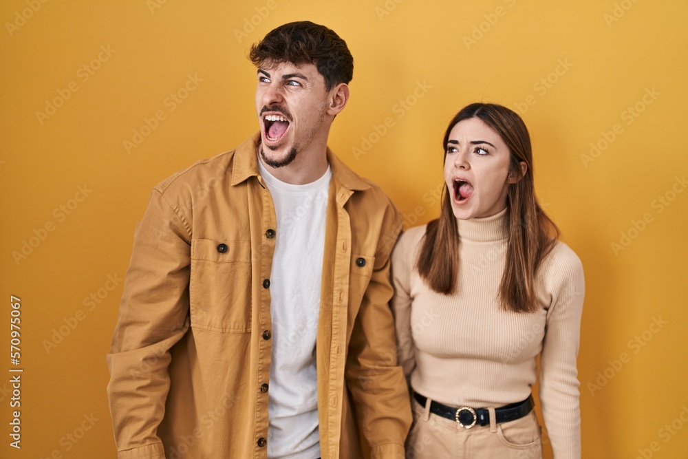 Sticker young hispanic couple standing over yellow background angry and mad screaming frustrated and furious