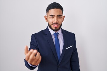 Young hispanic man wearing business suit and tie beckoning come here gesture with hand inviting welcoming happy and smiling