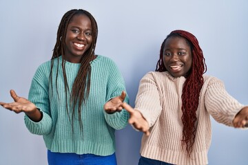 Two african woman standing over blue background smiling cheerful offering hands giving assistance and acceptance.