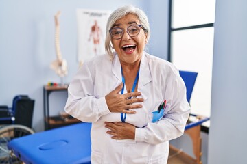 Middle age woman with grey hair working at pain recovery clinic smiling and laughing hard out loud because funny crazy joke with hands on body.